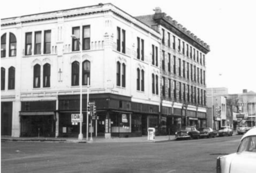 Black and white photo of a historic building corner with shops and parked cars on a city street.