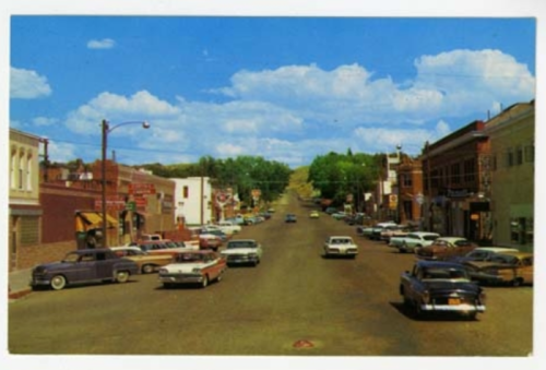 A vintage street scene with classic cars parked along a tree-lined road under a blue sky with fluffy clouds.