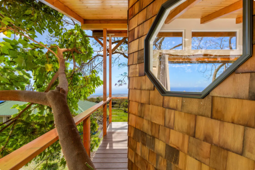 A wooden walkway leads to a cozy cabin with a large window, surrounded by greenery and a scenic view.