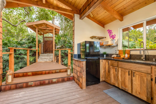 Cozy wooden kitchen with a black fridge, leading to a garden path surrounded by lush greenery.