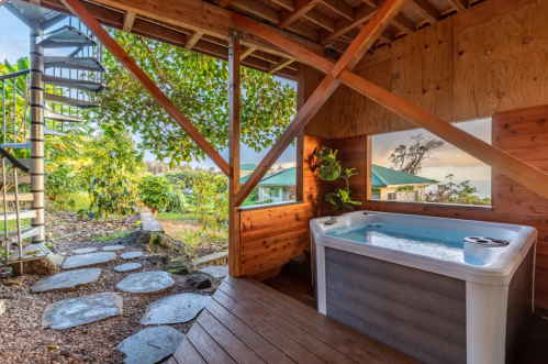 A wooden deck with a hot tub, surrounded by greenery and a spiral staircase leading to an upper level.
