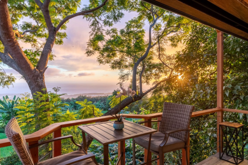A serene balcony view with a table and chairs, overlooking lush greenery and a sunset by the ocean.