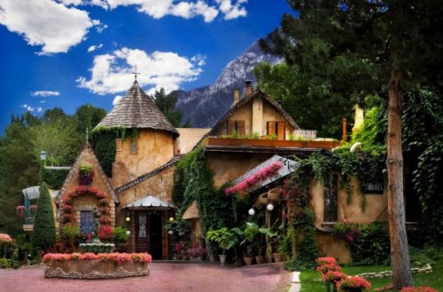 A charming, ivy-covered cottage with a turret, surrounded by vibrant flowers and tall trees against a mountain backdrop.