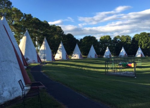 A row of white teepees on a grassy field under a blue sky with scattered clouds.