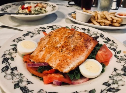 A plate of grilled salmon on a bed of greens, topped with tomato, onion, and sliced hard-boiled eggs, with fries in the background.