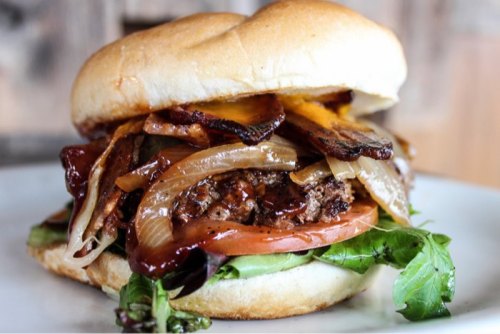 A juicy burger with melted cheese, grilled onions, bacon, tomato, and lettuce on a soft bun, served on a plate.