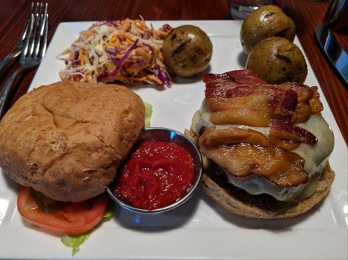 A plate featuring a bacon cheeseburger, coleslaw, and three roasted potatoes with a side of ketchup.