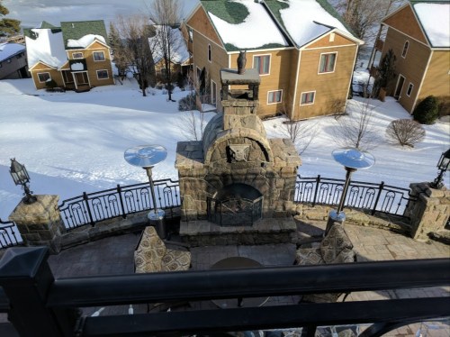 A stone outdoor fireplace surrounded by patio furniture, with snow-covered ground and houses in the background.