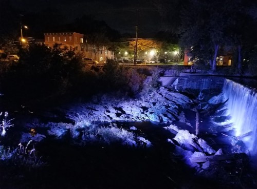 A nighttime scene featuring a waterfall illuminated by blue lights, with a building and trees in the background.