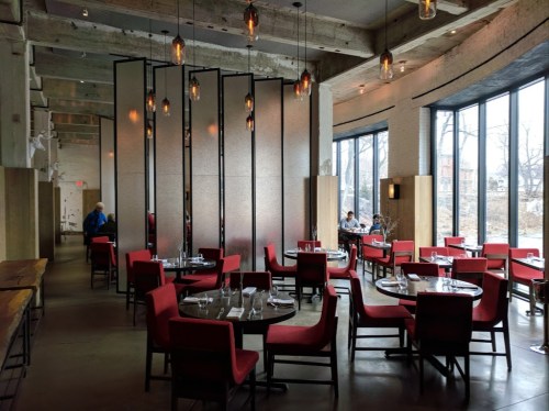 Modern restaurant interior with red chairs, round tables, and large windows, featuring a minimalist design and warm lighting.
