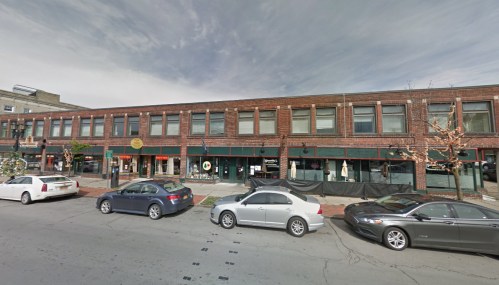A street view of a brick building with shops and parked cars under a cloudy sky.