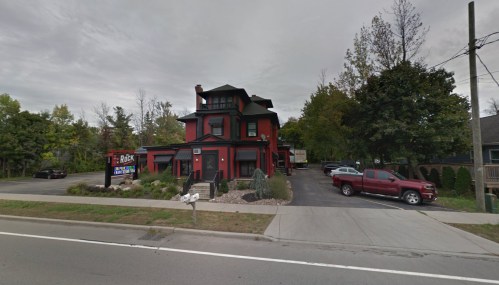 A large red house with a black roof, surrounded by trees, parked cars, and a sign in front, on a cloudy day.