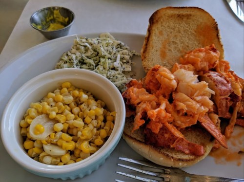 A plate featuring a lobster sandwich on bread, coleslaw, and a side of corn.