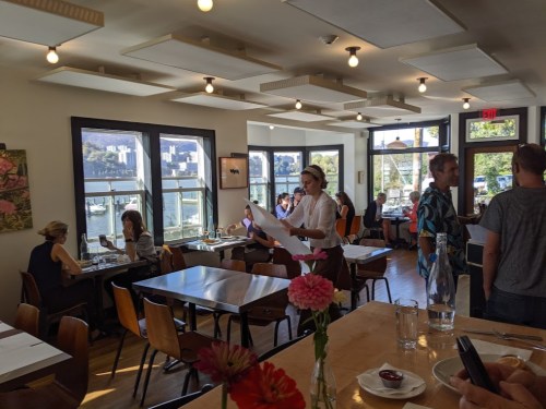 A busy restaurant interior with diners at tables, a server holding a menu, and large windows showing a view outside.