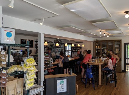 A cozy café interior with a wooden counter, patrons ordering, and shelves filled with snacks and drinks.