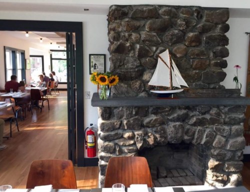 A cozy restaurant interior featuring a stone fireplace, a model sailboat, and sunflowers on the mantel.