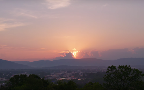 A serene sunset over mountains, with soft pink and orange hues illuminating the sky and clouds.