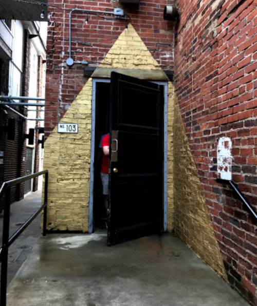 A black door with a golden triangle design on a brick wall, partially open, revealing a person inside.