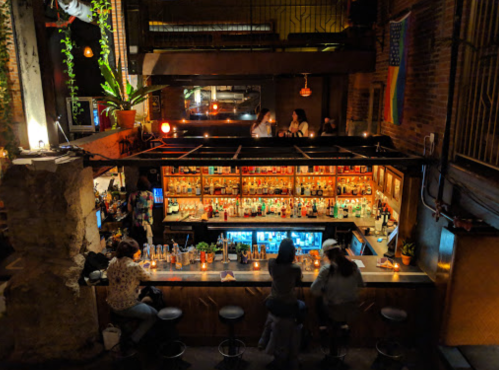 A vibrant bar scene with colorful bottles, patrons seated at the counter, and plants decorating the space.
