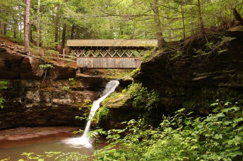 A wooden covered bridge spans a rocky area with a waterfall, surrounded by lush green trees and foliage.