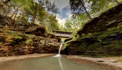 A serene waterfall cascades into a pool, framed by lush greenery and a rustic covered bridge above.