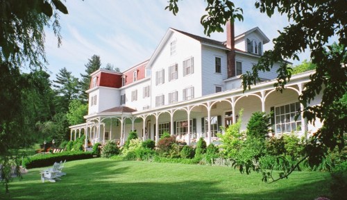 A large white house with a red roof, surrounded by greenery and a porch, set against a blue sky.