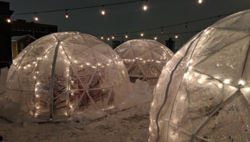 Three illuminated igloos on a snowy rooftop, surrounded by string lights against a dark sky.