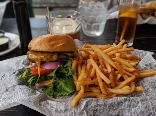 A juicy cheeseburger with lettuce, tomato, and onion, served with a side of crispy French fries on newspaper.