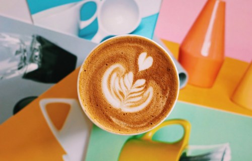 A top view of a latte with heart and leaf art, surrounded by colorful mugs and geometric shapes.
