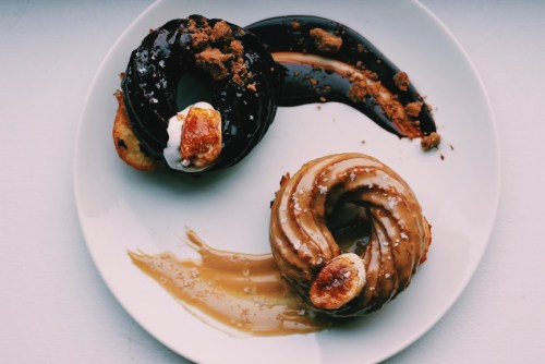 A plate with two donuts: one chocolate-glazed with crumbs, the other caramel-glazed with a piece of banana on top.
