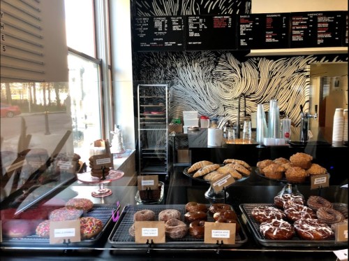 A display case filled with various donuts and pastries, with a menu board and artistic wall design in the background.