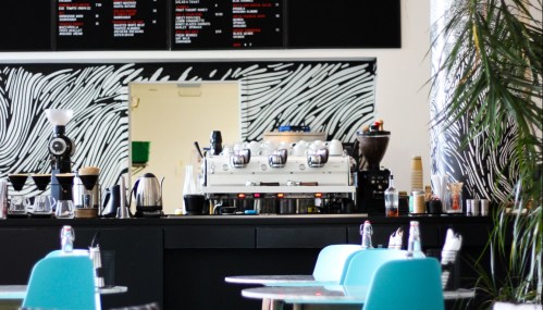 A modern café interior featuring a coffee machine, black counter, and turquoise chairs against a patterned wall.
