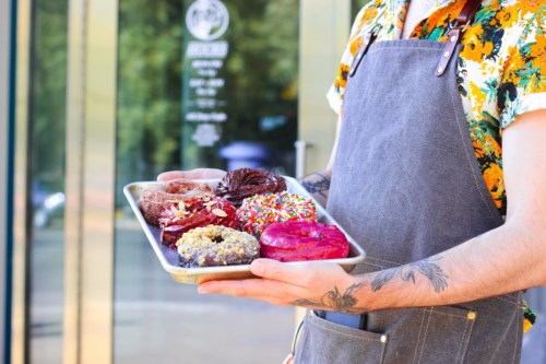 A person in a floral shirt and apron holds a tray of colorful donuts outside a shop.