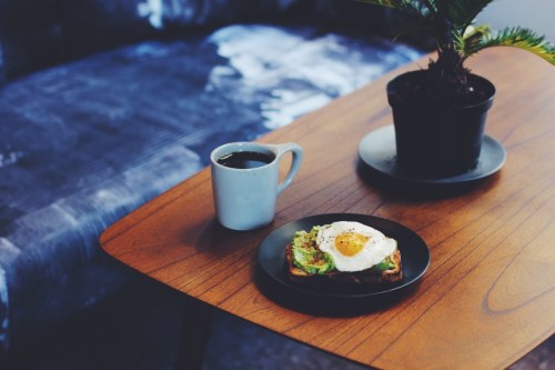 A wooden table with a plate of avocado toast topped with a fried egg, a cup of coffee, and a potted plant.