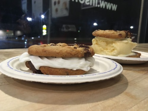 Two cookie sandwiches on paper plates, one filled with white cream and the other with ice cream, set on a wooden table.