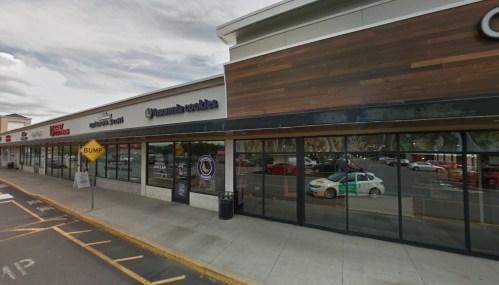 Exterior view of a shopping plaza featuring a cookie shop and other storefronts, with parking spaces visible.
