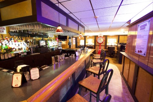 A cozy bar interior with a wooden counter, shelves of liquor, and vintage decor, featuring a clock and seating.