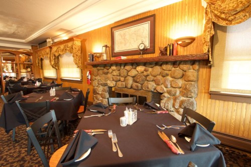 Cozy restaurant interior with stone fireplace, wooden walls, and neatly set tables with black tablecloths and utensils.
