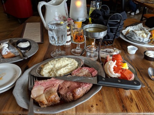 A table set with a plate of prime rib, mashed potatoes, lobster, drinks, and a candle in a cozy restaurant setting.