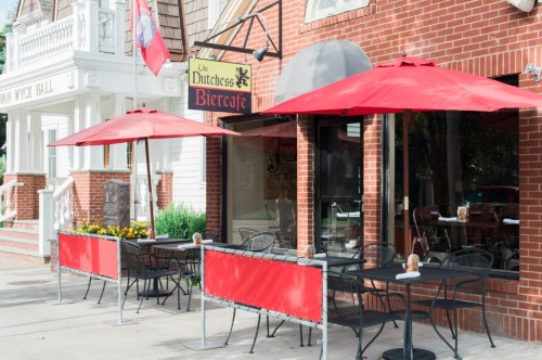 Outdoor seating area of a café with red umbrellas, brick walls, and a sign reading "Cafe Dutchess Biercafe."