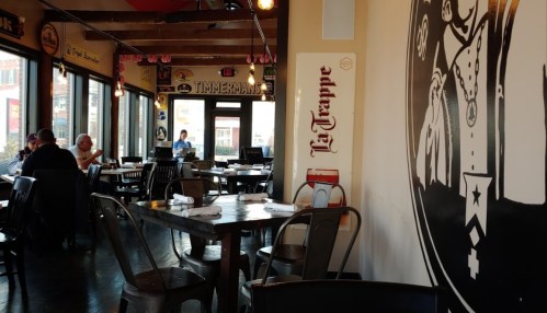 Interior of a restaurant with wooden tables, chairs, and a mural on the wall; patrons are seated at various tables.