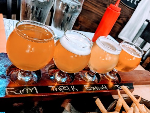 A wooden flight tray holds four small glasses of different beers, with a red condiment bottle in the background.