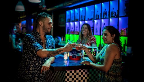 A man and woman clink glasses at a vibrant bar, with colorful lights and other patrons in the background.