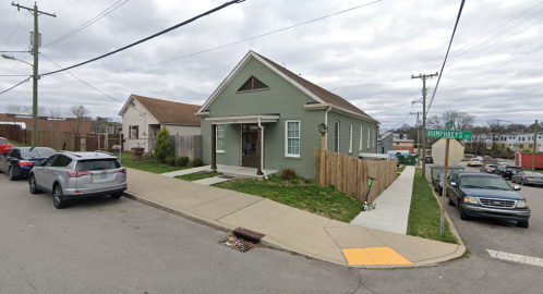 A green house on a corner lot with a sidewalk, parked cars, and cloudy skies in the background.