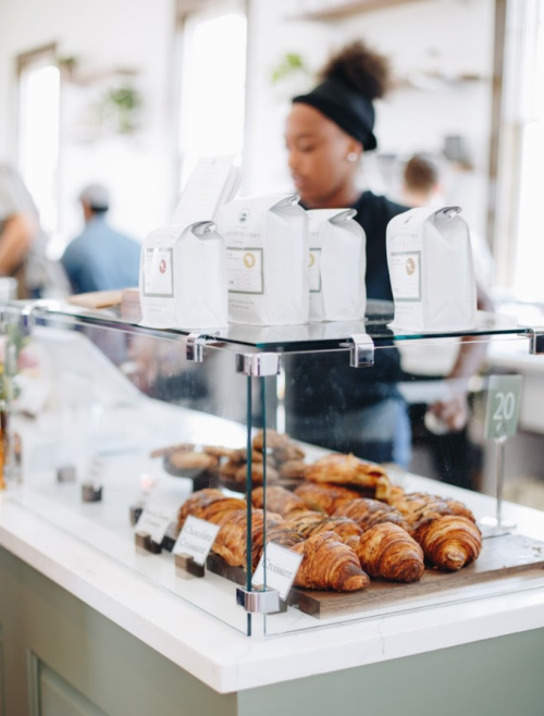 A display case with croissants and coffee bags, with a barista in the background at a cozy café.