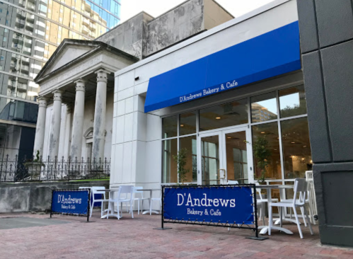 Exterior of D'Andrews Bakery & Café featuring outdoor seating and a blue awning, set against a modern building backdrop.