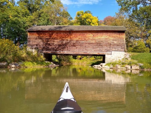 A wooden covered bridge reflected in calm water, surrounded by trees with autumn foliage under a clear blue sky.