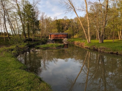 A serene landscape featuring a wooden cabin by a calm stream, surrounded by lush greenery and trees reflecting in the water.