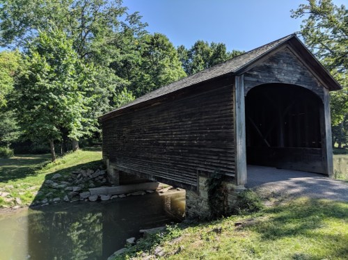 A wooden covered bridge spans a calm pond, surrounded by lush green trees under a clear blue sky.