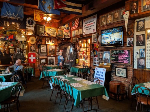 Cozy restaurant interior with green tables, framed photos on brick walls, and a TV showing a basketball game.